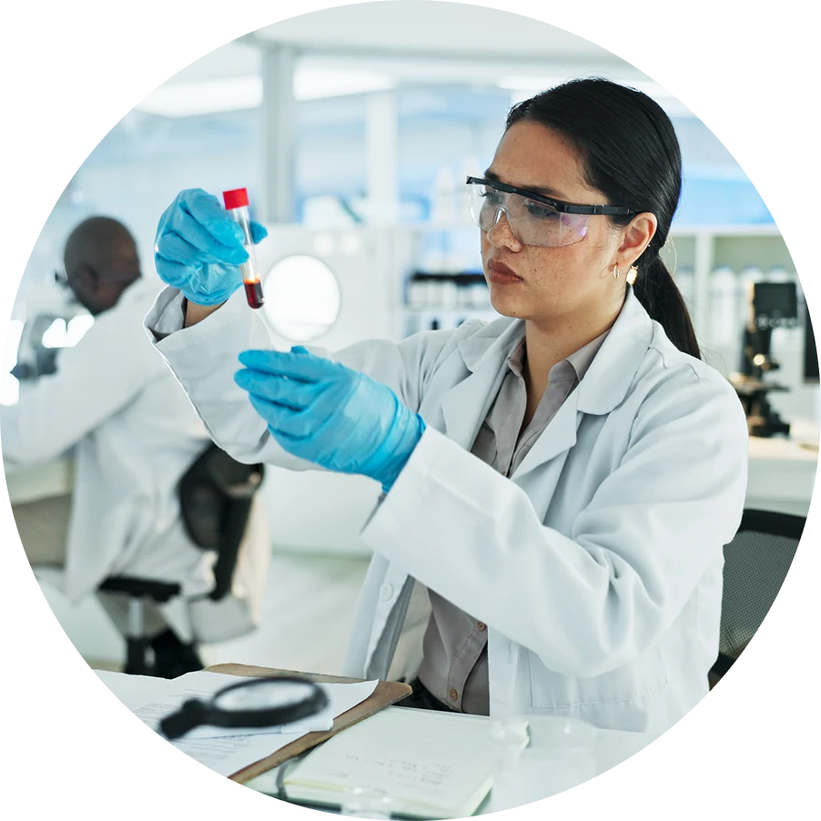 Female scientist in safety goggles and blue gloves holds a test tube with red liquid in a bright laboratory.